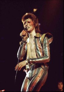 David Bowie performs on stage in a glitter striped jumpsuit.