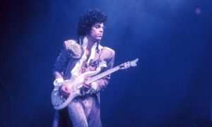 Prince performs on stage wearing a sequinned outfit and playing an electric guitar.